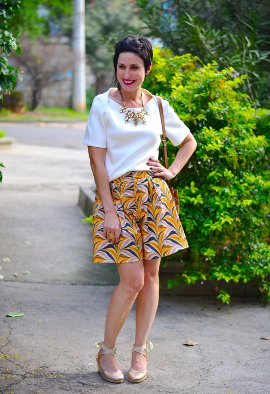 short-saia com estampa de folhagens azuis, rosas e brancas em funco amarelo, blusa de neoprene armada branca, colar dourado de triângulos e espadrilhe dourada