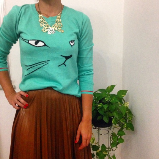 blusa de manga comprida verde água com olhos e focinho de gato, saia marrom plissada de couro falso e colar de arabescos dourado