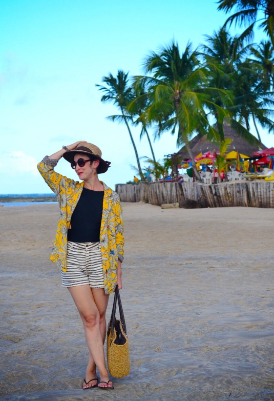 blazer amarelo largo com estampa de coqueiros preto e branco, maiô preto, chinelo e short listrado azul marinho e branco