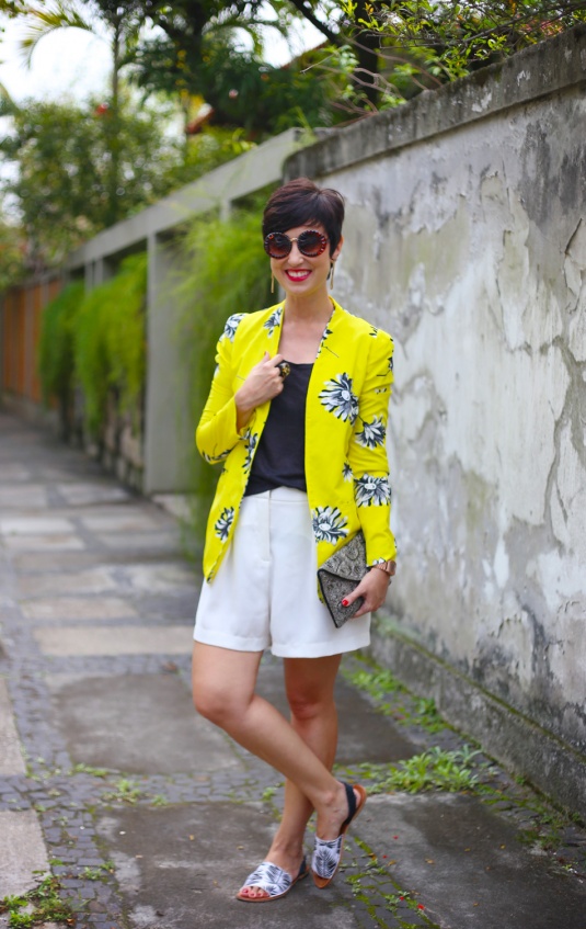 blazer amarelo com estampa de margaridas em preto e branco, bermuda branca soltinha, sandália branca com estampa de folhas de palmeiras em preto, top preto e carteira d emão bordada cinza