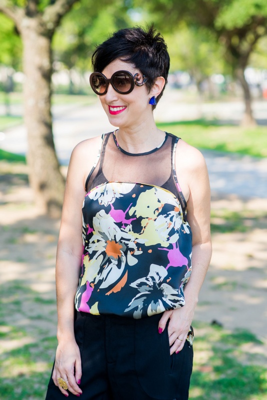 regata de seda com transparência preta em cima e estampa floral de fundo escuro, rosa, branco e laranja, calça preta curta com alfinetes e laterais abertas, scarpin verde metalizado, colete de couro preto