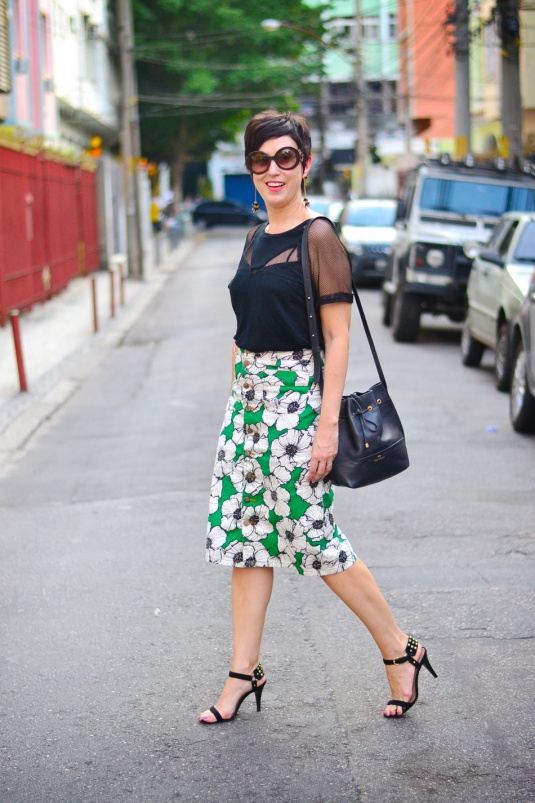 saia de brim com botões na frente e estampa de floresm em preto e branca no fundo verde aberto, comprida até o joelho; blusa preta de mangas curtas com detalhes telados dando transparência; sandália de duas tiras preta e bolsa bucket bag preta