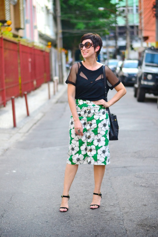 saia de brim com botões na frente e estampa de floresm em preto e branca no fundo verde aberto, comprida até o joelho; blusa preta de mangas curtas com detalhes telados dando transparência; sandália de duas tiras preta e bolsa bucket preta