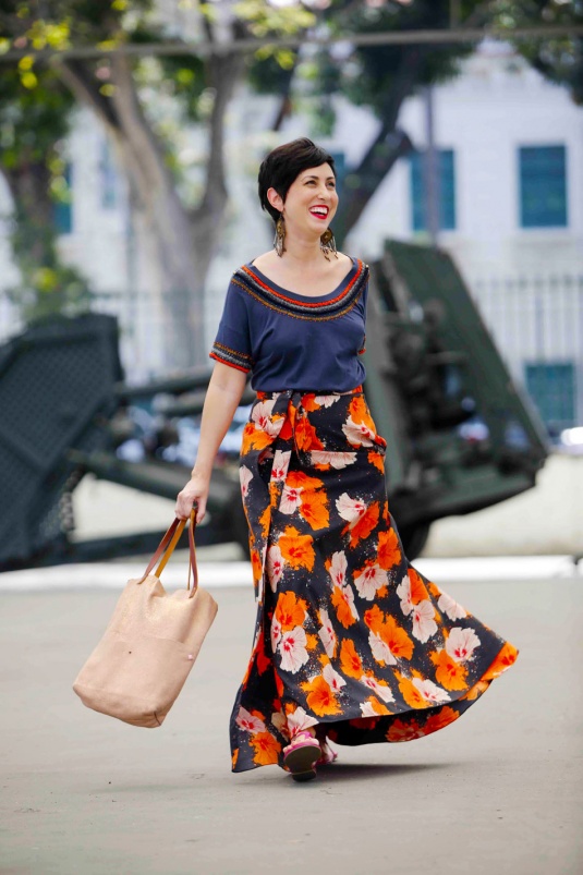 saia longa estruturada com fundo preto e estampa de flores tropicais laranjas, brancas e rosas, camiseta azul escura com miçangas laranjas, azuis claras e douradas nas mangas e gola, rasteira rosa, bolsa metalizada rosé