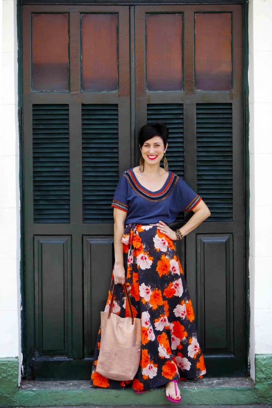 saia longa estruturada com fundo preto e estampa de flores tropicais laranjas, brancas e rosas, camiseta azul escura com miçangas laranjas, azuis claras e douradas nas mangas e gola, rasteira rosa, bolsa metalizada rosé