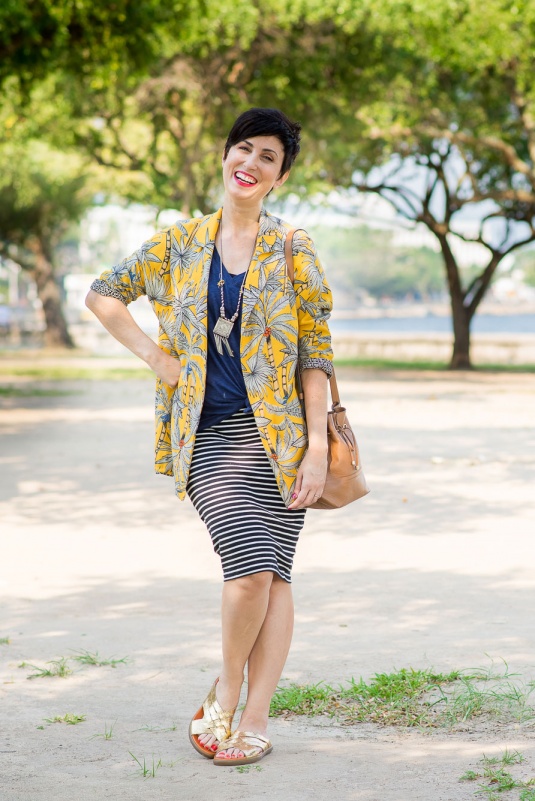 t-shirt azul escura com saia lápis de malha listrada pb, sandália chinelo de tiras cruzadas douradas metalizadas, bolsa saco caramelo, colar comprido, blazer tipo quimono amarelo com estampa de coqueiros brancas, azuis e vermelhas