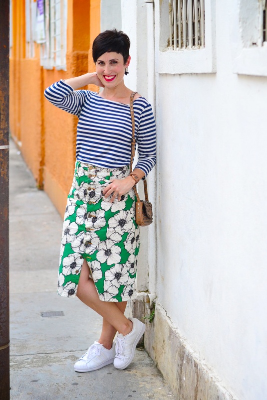 blusa de malha com mangas listrada azul e branca, saia mídi reta de brim com estampa verde com flores brancas e pretas e botões frontais, tênis adidas branco