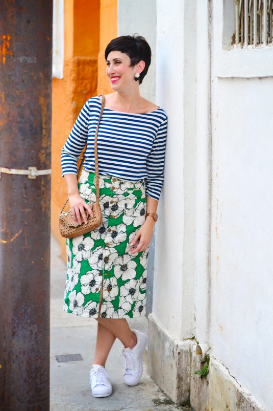blusa de malha com mangas listrada azul e branca, saia mídi reta de brim com estampa verde com flores brancas e pretas e botões frontais, tênis adidas branco